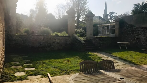 Une cour avec un chemin en briques, une table en bois entourée de chaises, un mur en pierre et une vue sur le clocher d’une église.