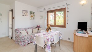Una habitación con un sofá floral, una mesa de comedor con mantel floreado, un televisor sobre un soporte de madera y una ventana con vistas a la vegetación.