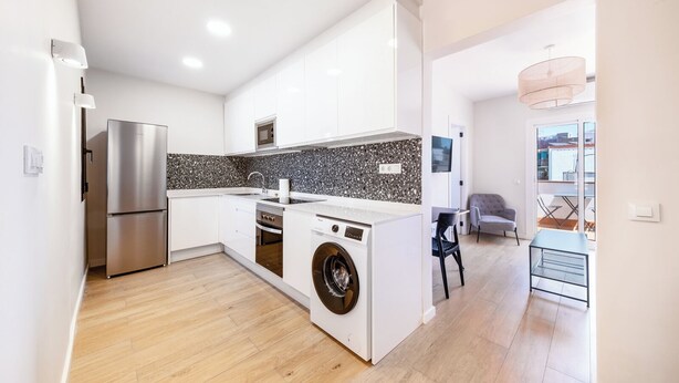 A modern kitchen with a stainless steel refrigerator, a washing machine, and a dining area with a table and chairs. - Recently refurbished 3 bedroom apartment with terrace (Barcelona)