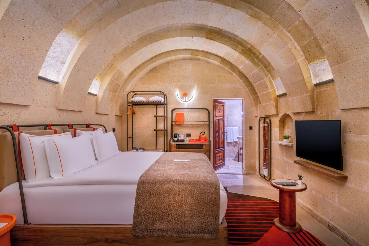 A hotel room with a bed, a desk, a TV, and an arched stone ceiling.