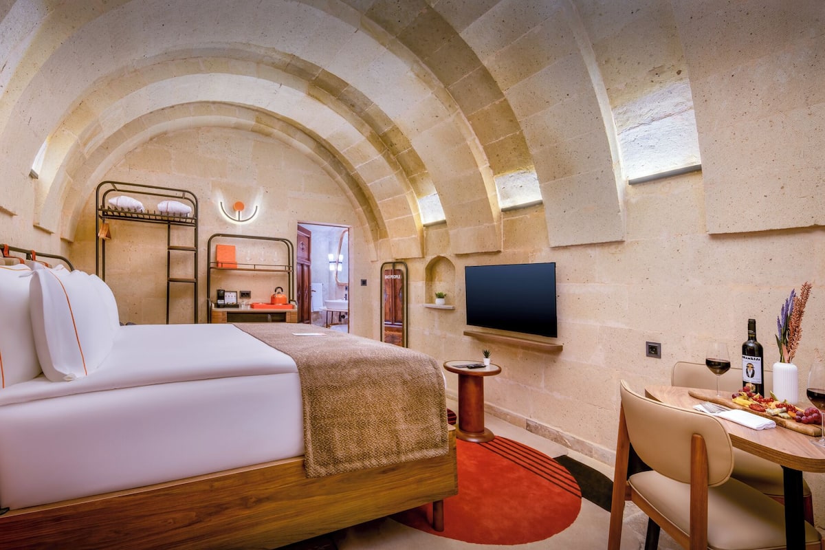 Habitación de hotel con cama, escritorio y silla, televisor y mesa de comedor con copas de vino.
