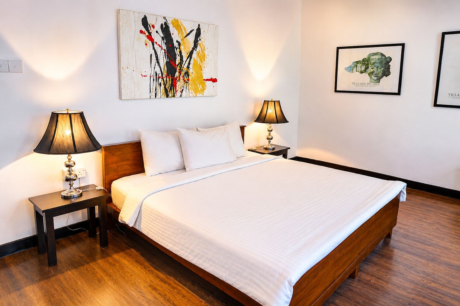 A hotel room with a large bed, two bedside tables with lamps, a wall-mounted abstract painting, and two framed pictures.