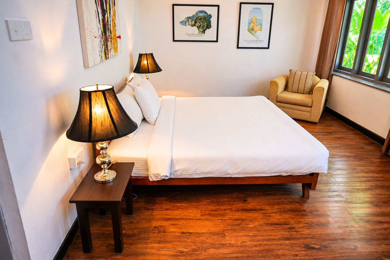 A bedroom with a wooden bed, two bedside lamps, a chair, and framed artwork on the wall.