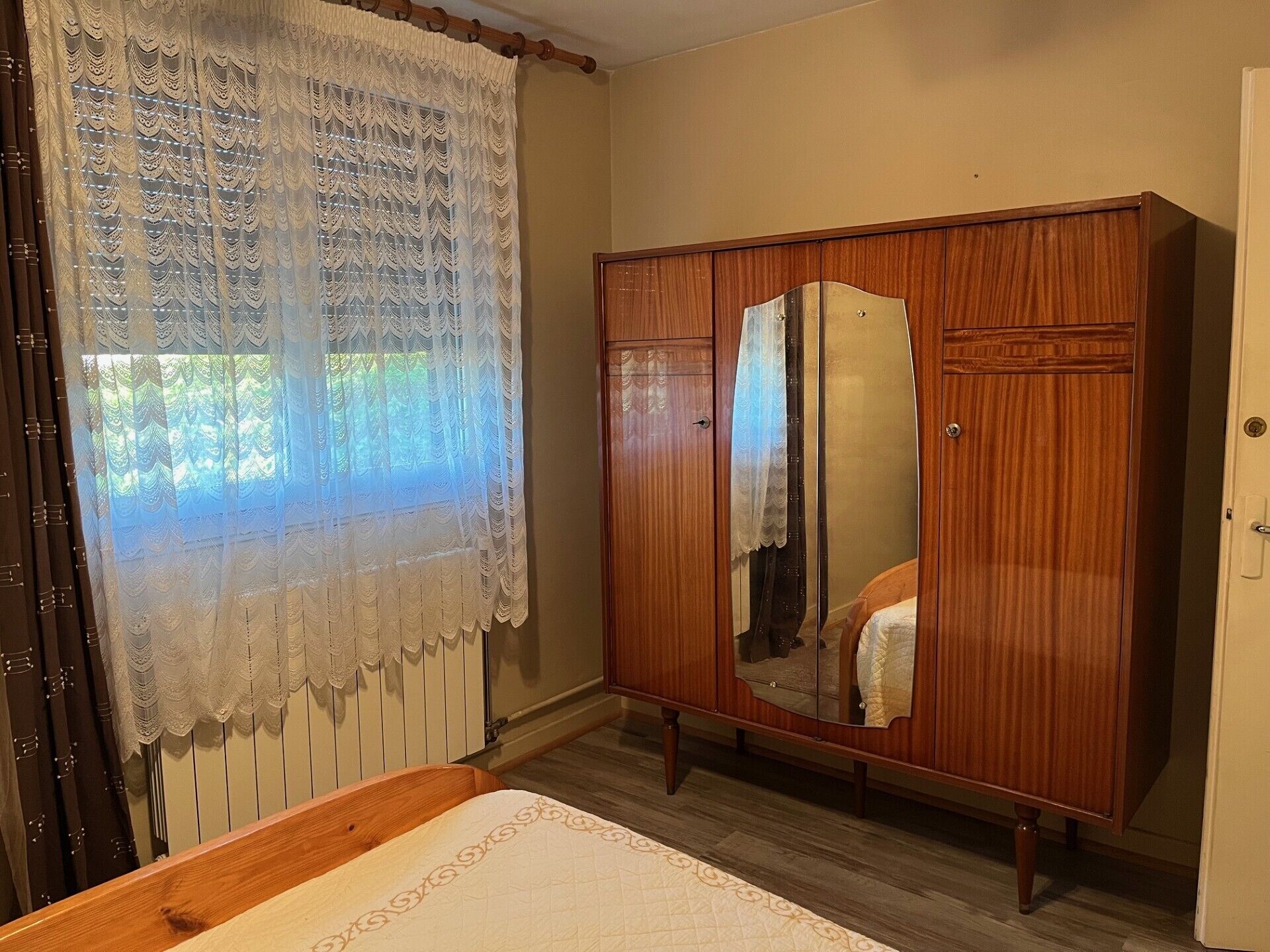 Un dormitorio con un armario de madera, una cama con colcha y una ventana con cortinas de encaje.