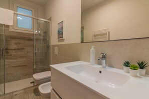 Un baño moderno con cabina de ducha de vidrio, un lavamanos blanco con dispensador de jabón y un inodoro.