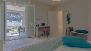 A bedroom with a bed, a desk, and a chair, a suitcase, and a view of a body of water.
