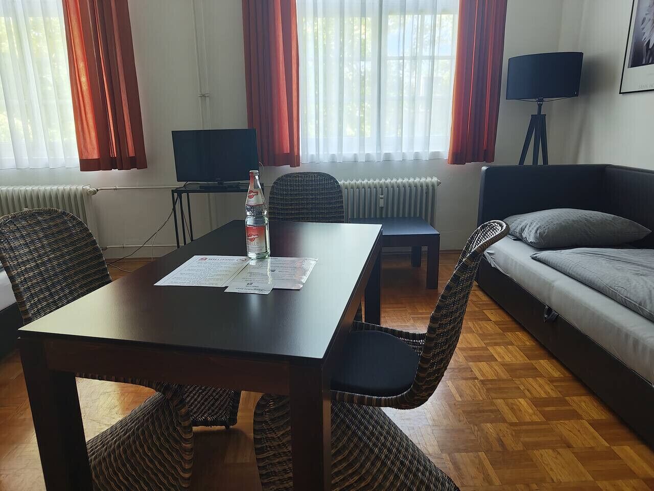 A room with a wooden table, wicker chairs, a sofa, and a bed. There is a radiator, a lamp, and a television on a stand.