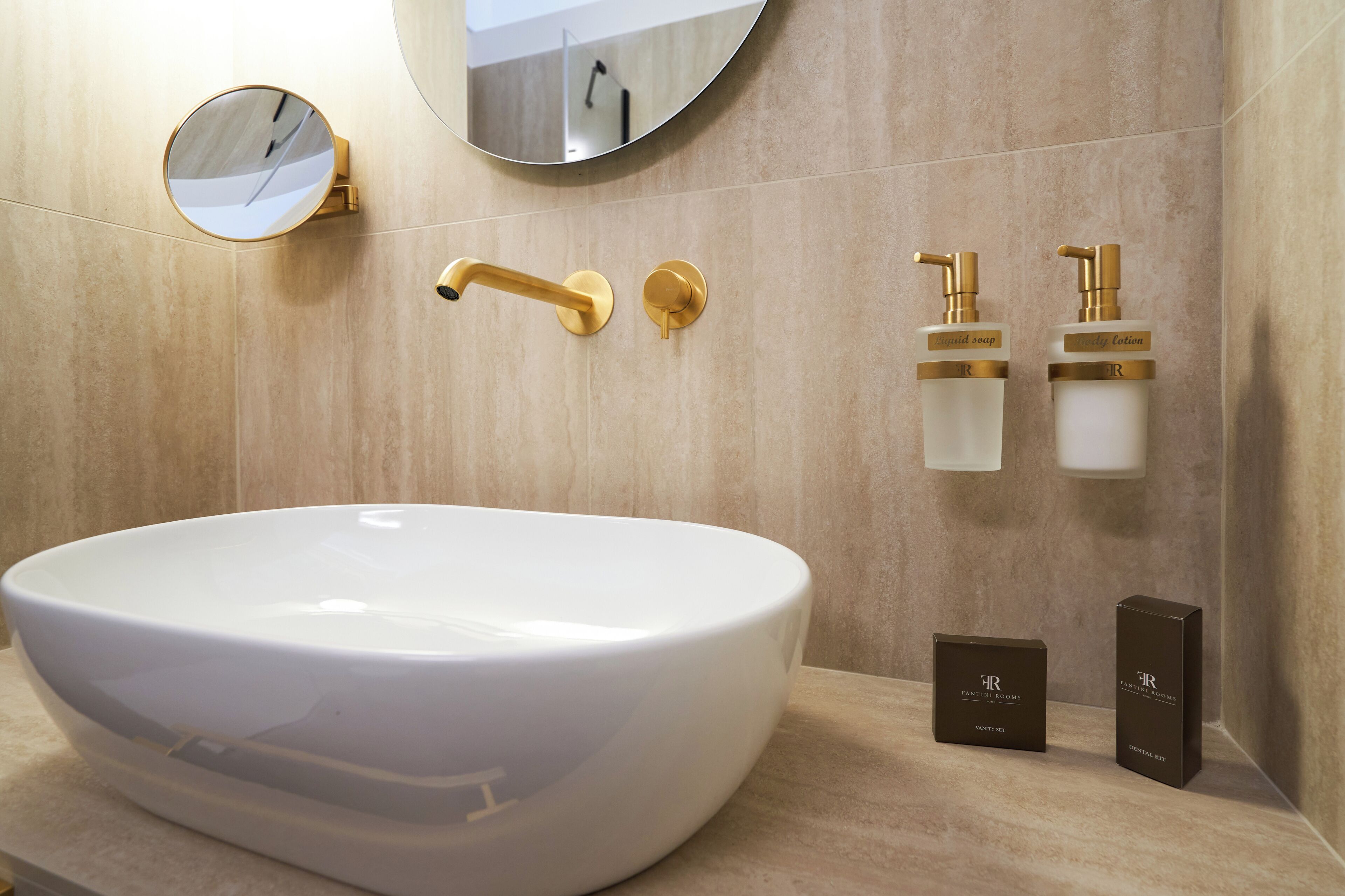 Un baño con un lavabo grande blanco, accesorios dorados, un espejo redondo y dos dispensadores de jabón.