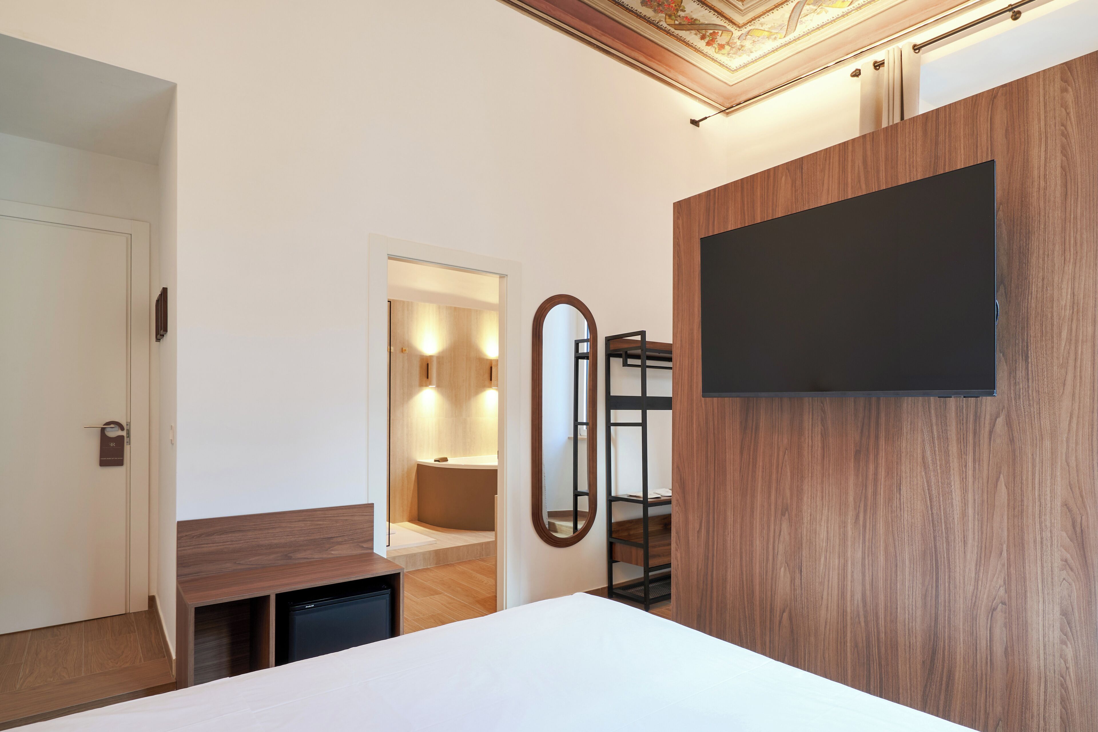 A hotel room with a bed, a small desk, a mirror, a TV mounted on the wall, and a bathroom visible through an open door.