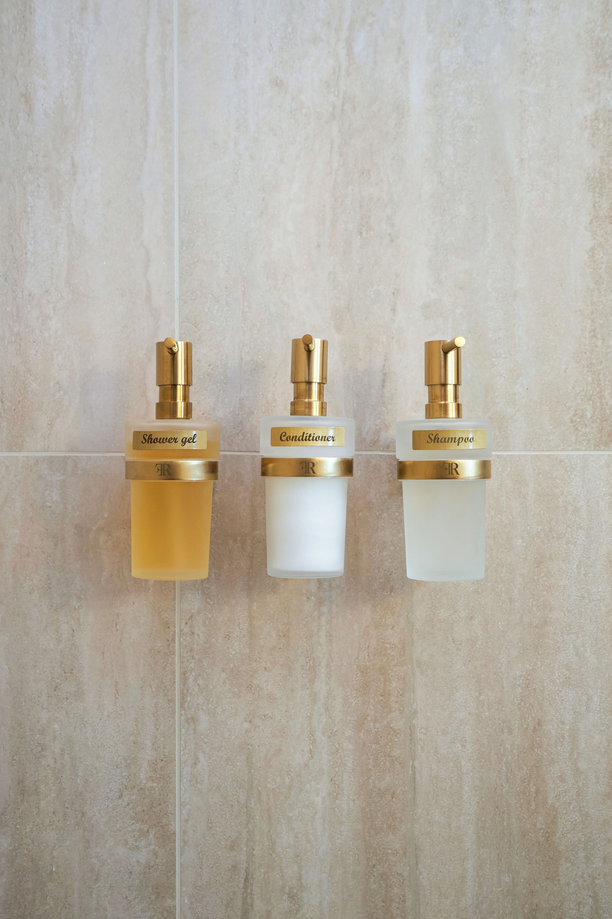 Trois flacons de produits de toilette, étiquetés « gel douche », « après-shampoing » et « shampoing », sont fixés à un mur recouvert de carreaux.