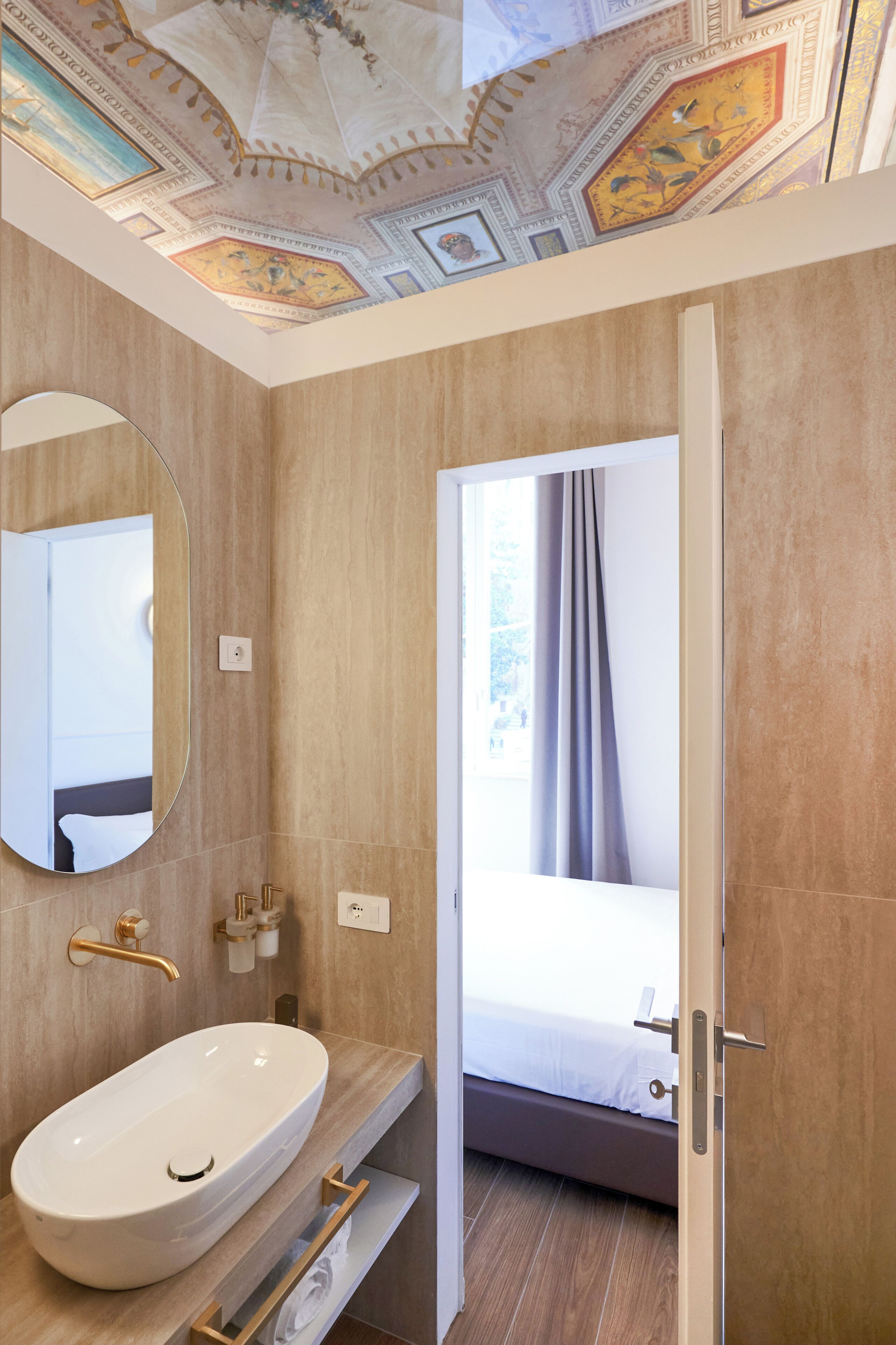 A modern bathroom with a large oval sink, a mirror, and a door leading to a bedroom with a bed and curtains.