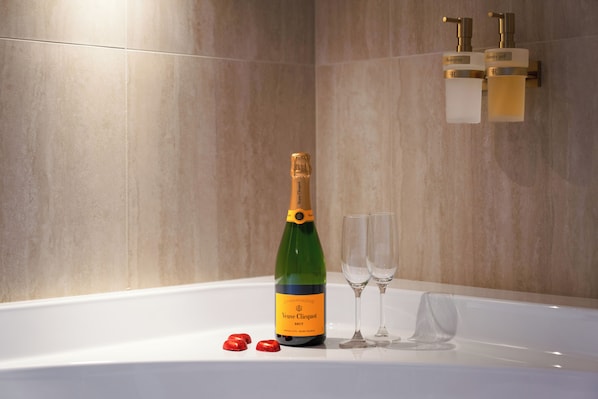 A champagne bottle and two glasses on a white surface, with a tiled wall and a wall-mounted dispenser in the background. - Fantini Rooms- Fori Imperiali (Rome)