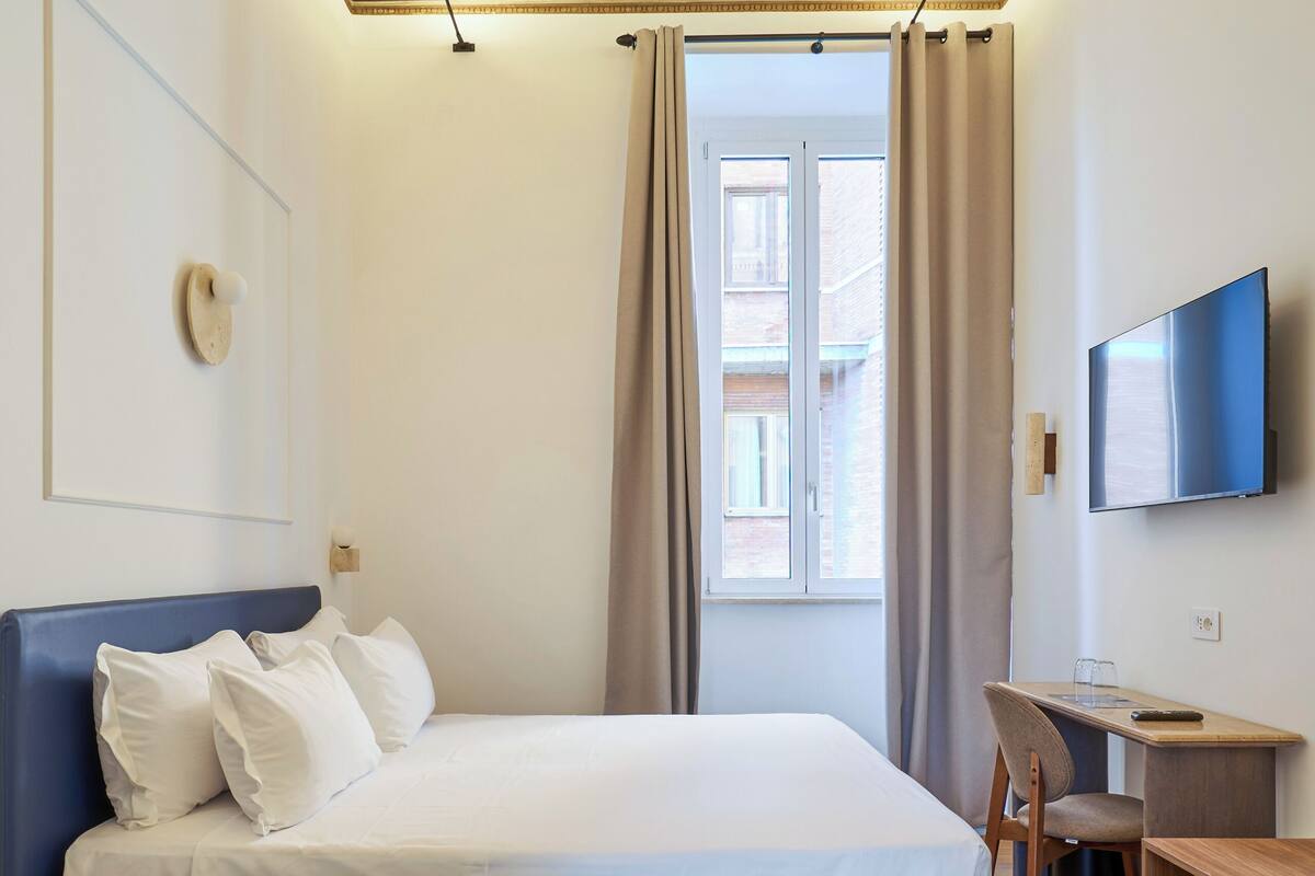 A hotel room with a bed, a desk, a television, a window with curtains, and a decorative ceiling.