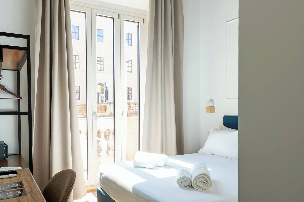 A hotel room with a bed, a desk, a chair, a window with curtains, and a ceiling with a decorative design.