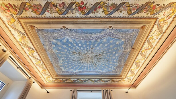 A room with a detailed ceiling fresco, a window with curtains, and a wooden beam structure. - Fantini Rooms- Fori Imperiali (Rome)