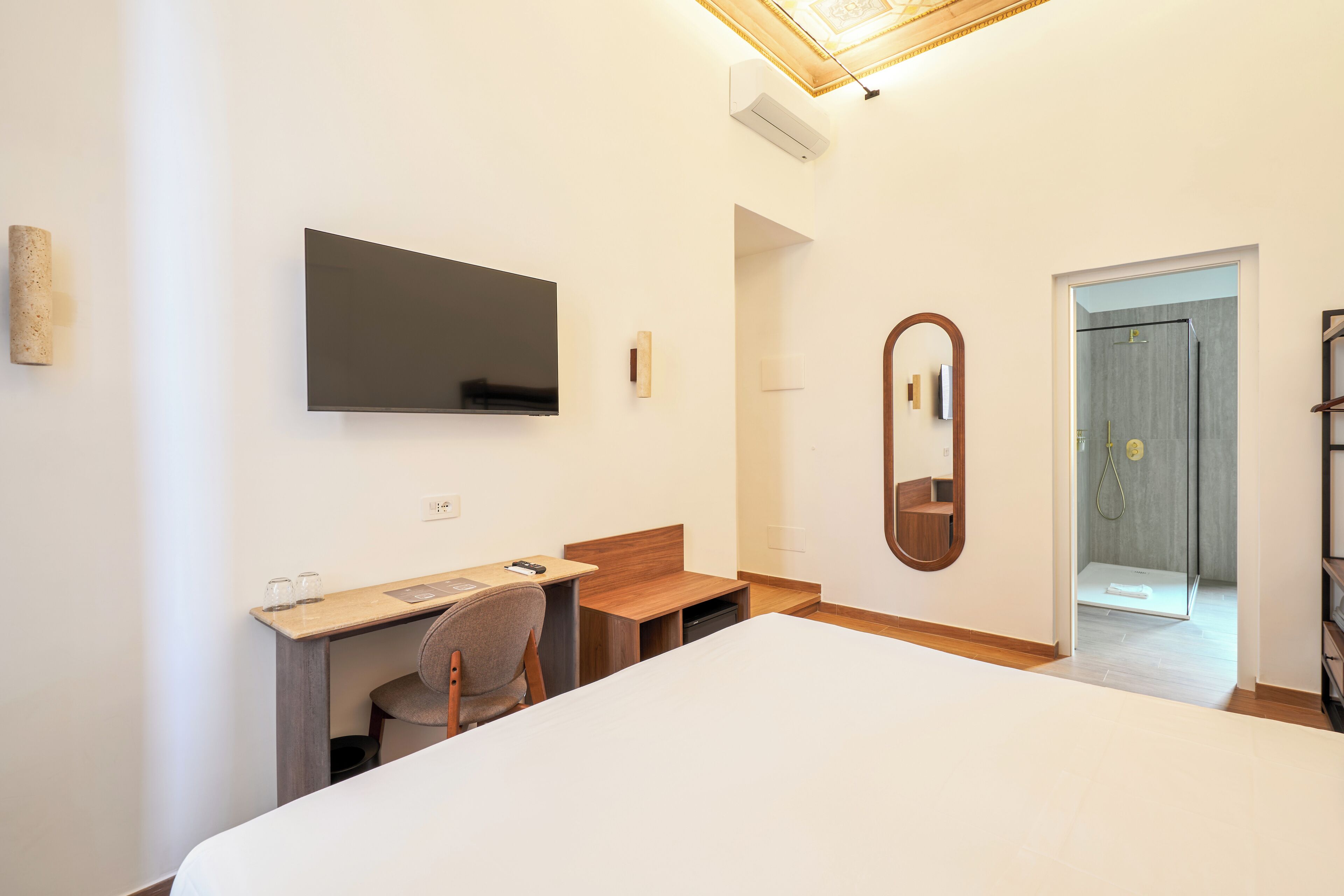 A hotel room with a bed, desk, chair, TV, mirror, and a door leading to a shower area.
