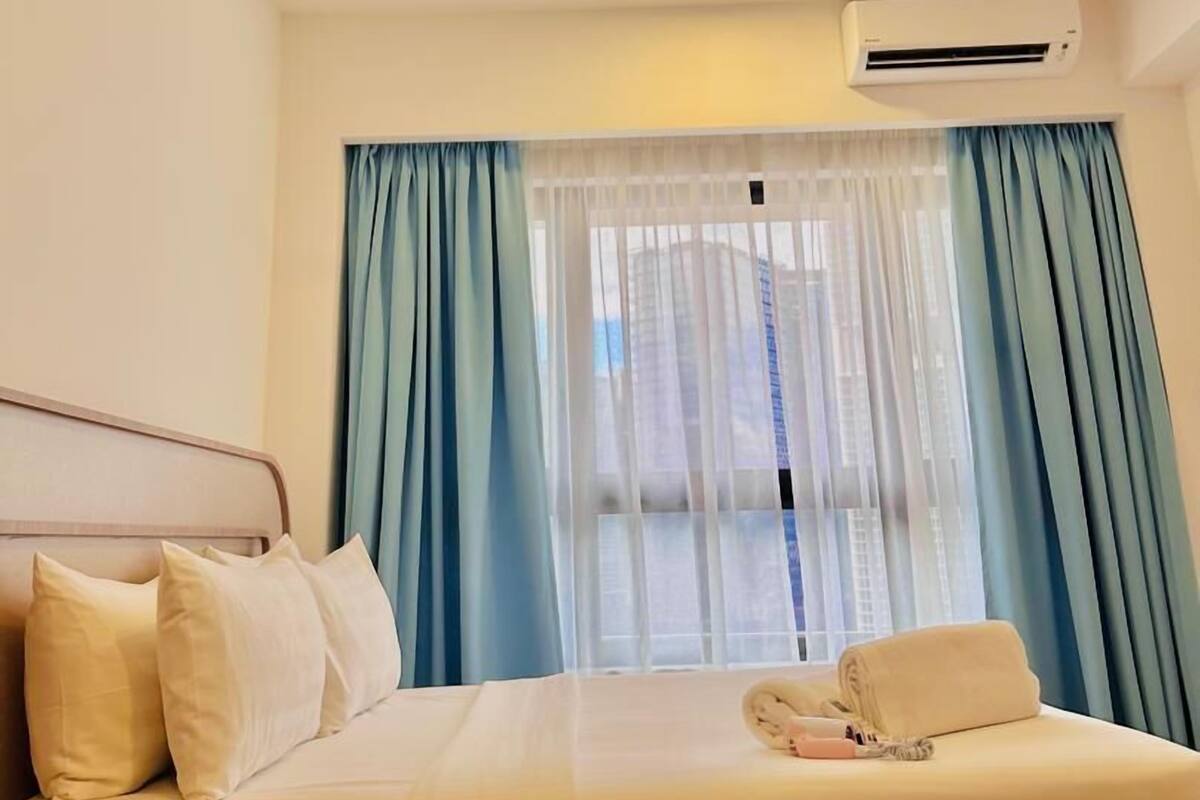 Une chambre d’hôtel avec un lit, un ventilateur de plafond, un appareil de climatisation et une fenêtre avec des rideaux bleus.
