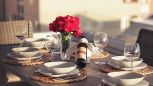 Una mesa de comedor dispuesta con platos, fuentes y copas de vino blancos, una botella de vino y un florero con flores rojas.