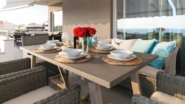 Un área de comedor al aire libre con mobiliario de mimbre, una mesa preparada para dos personas con platos y vasos blancos, y un jarrón con flores rojas.