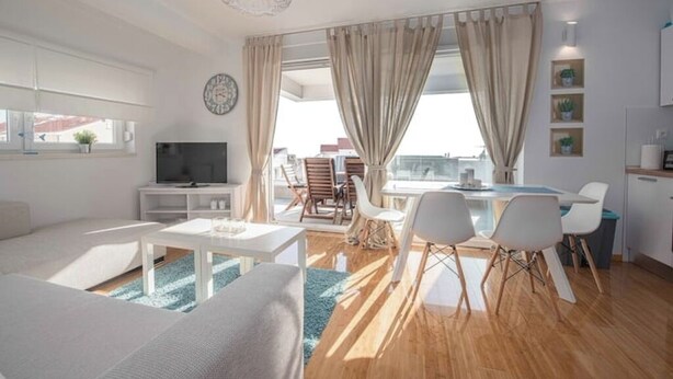 Un salon moderne comprenant un canapé, un téléviseur, un coin repas et de grandes fenêtres avec des rideaux.