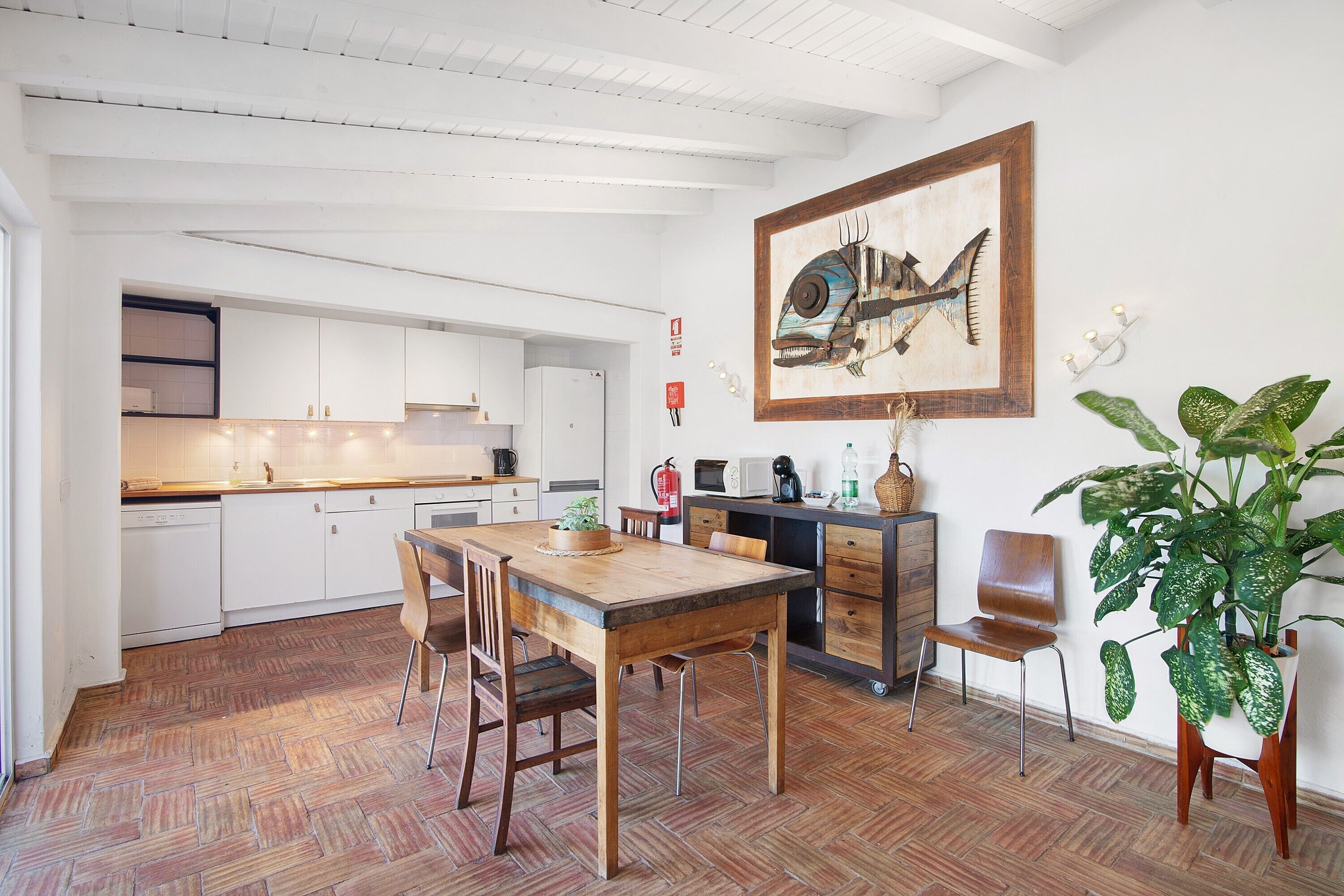 Eine Küche mit weißen Schränken, einem Holzespeisetisch, Stühlen, einem Blumentopf und einem gerahmten Kunstwerk an der Wand.