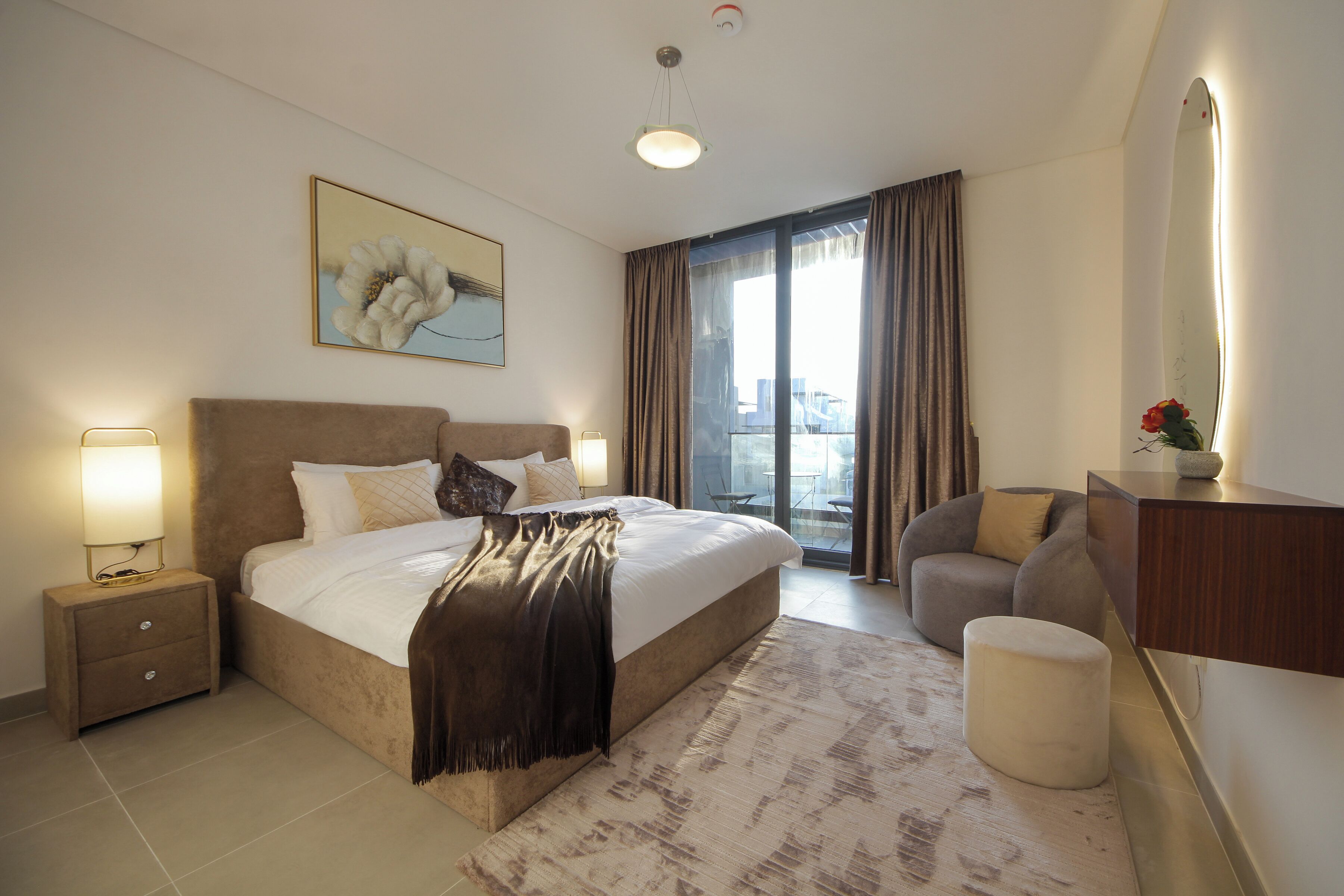 Ein modernes Hotelzimmer mit einem großen Bett, einem Nachttisch mit Lampe, einem Sessel, einem kleinen Hocker, einem großen Spiegel und Blick auf die Stadt durch das Fenster.