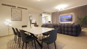 Een moderne woonkamer met een grote televisie, een eethoek met een witte tafel en zwarte stoelen, en een gezellige zithoek met een gestoffeerde bank.