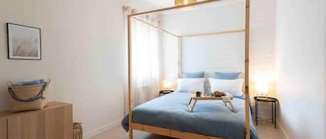 A bedroom with a four-poster bed, bedside tables, a bedside lamp, a framed picture on the wall, and a woven basket on the sideboard.