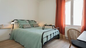 Une chambre à coucher comprenant un lit, une table de chevet, une chaise, une fenêtre avec des rideaux et un radiateur.