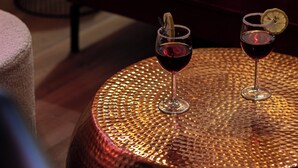 Une table ronde, dont le plateau est texturé, est posée sur un plancher en bois. On y voit deux verres de vin rouge et un cocktail.
