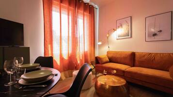 A living room with a sofa, a dining table with chairs, a television, and artwork on the wall.