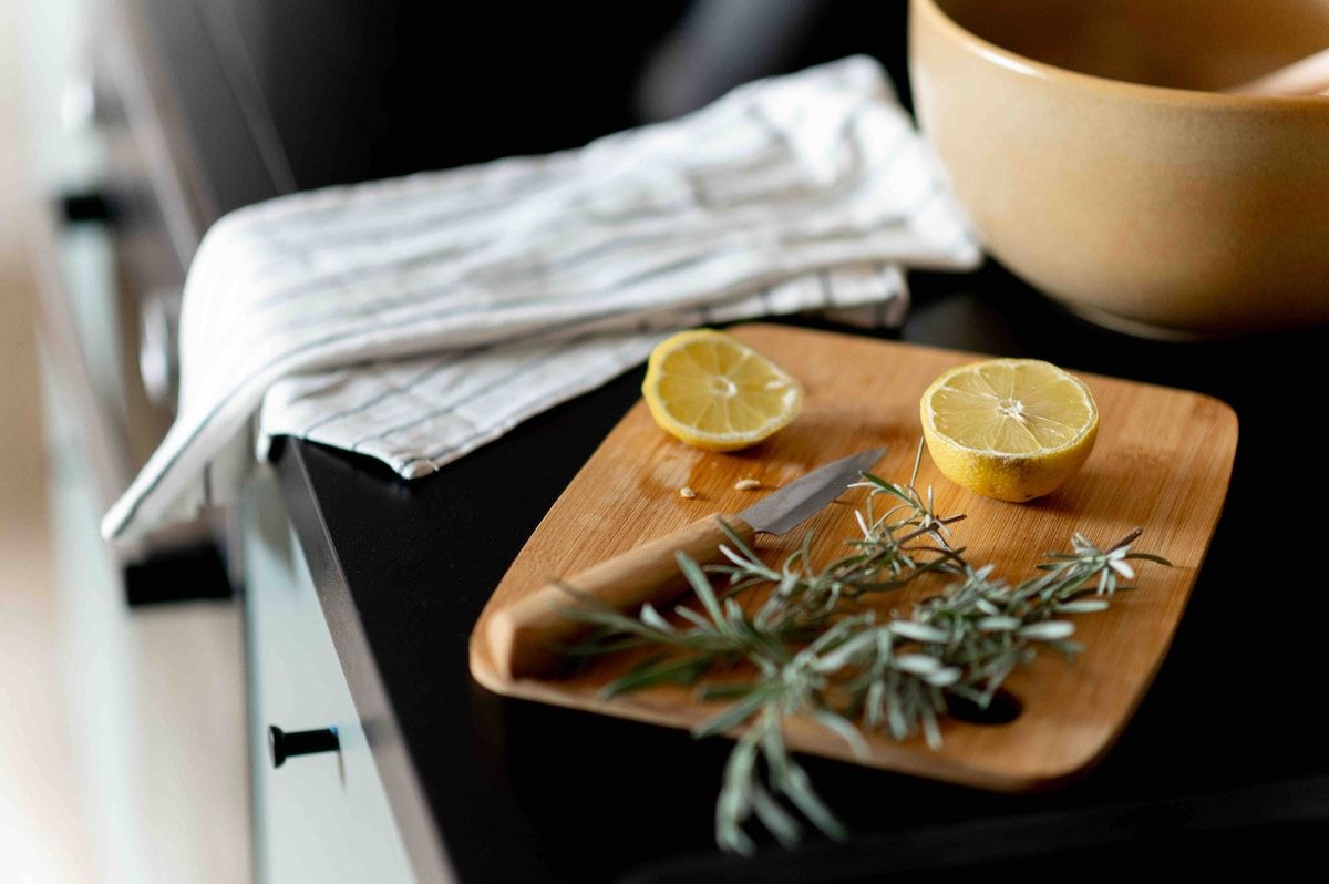 A wooden cutting board with two lemon halves and a knife, accompanied by a sprig of rosemary, on a kitchen counter.