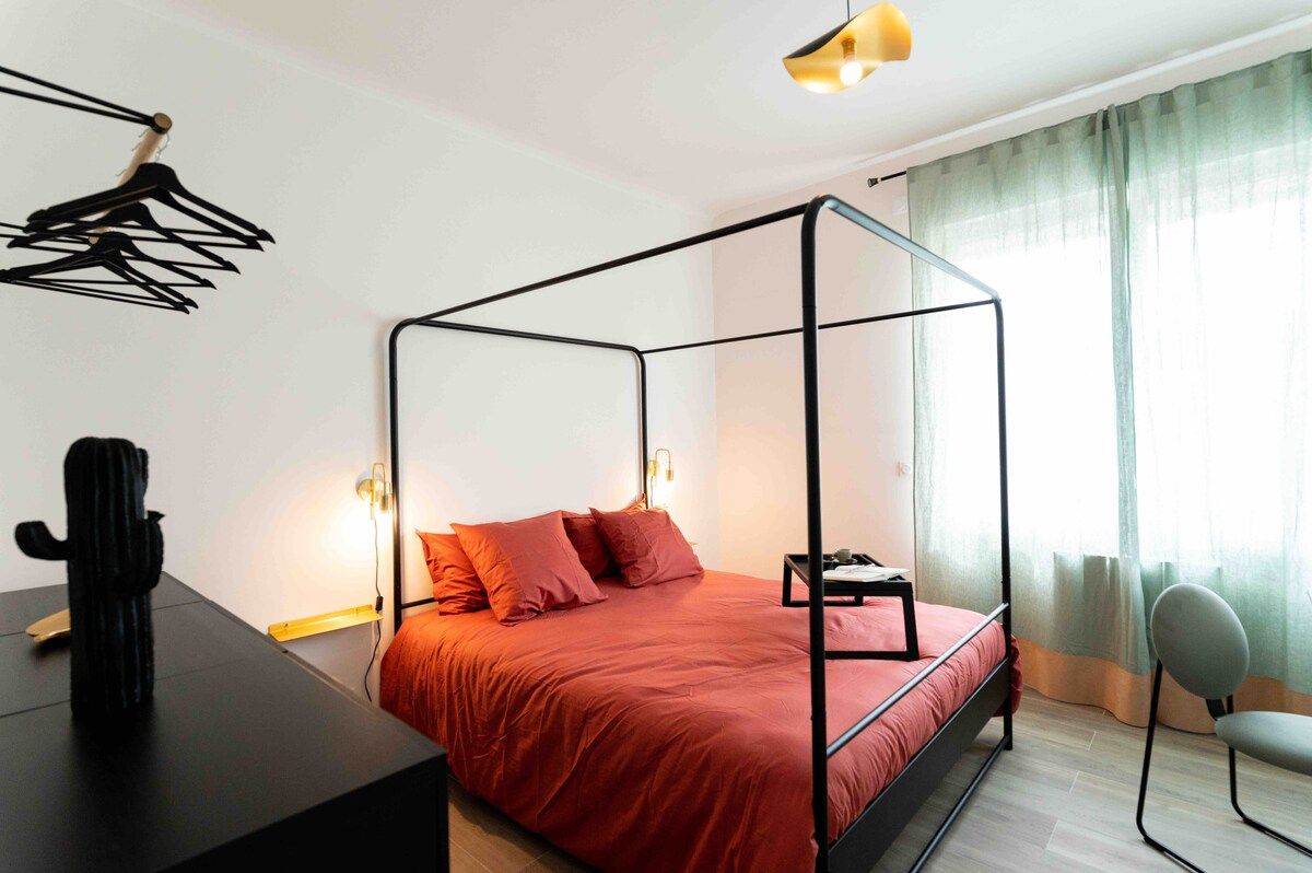 A room with a four-poster bed, a desk with a chair, a cactus, and a window with curtains.