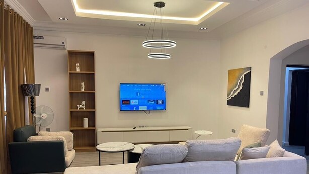 A modern living room with a sectional sofa, a flat-screen TV, a small round table, and a bookshelf. - Decorum Apartments (Abuja)
