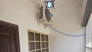 A room with a wooden door, a window with a grid pattern, two wicker chairs, a hanging light fixture, and an air conditioning unit. - Decorum Apartments (Abuja)