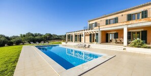 Une piscine avec des chaises longues, un patio couvert et un bâtiment doté de grandes fenêtres et de volets.