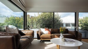 Una sala de estar moderna con sofás de cuero, una mesa de centro blanca y vistas a la vegetación y un edificio a través de amplios ventanales.