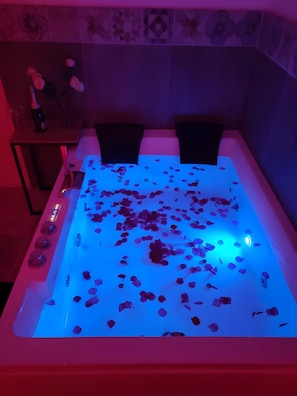 Une baignoire remplie de pétales de rose, éclairée d’une lumière bleue, avec une petite table et une bouteille à côté.
