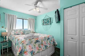 Un dormitorio con cama, ventilador de techo, ventana con cortinas, televisor montado en la pared y una puerta.