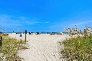 Un sendero arenoso que lleva al océano, con sombrillas y gente en la playa a lo lejos.
