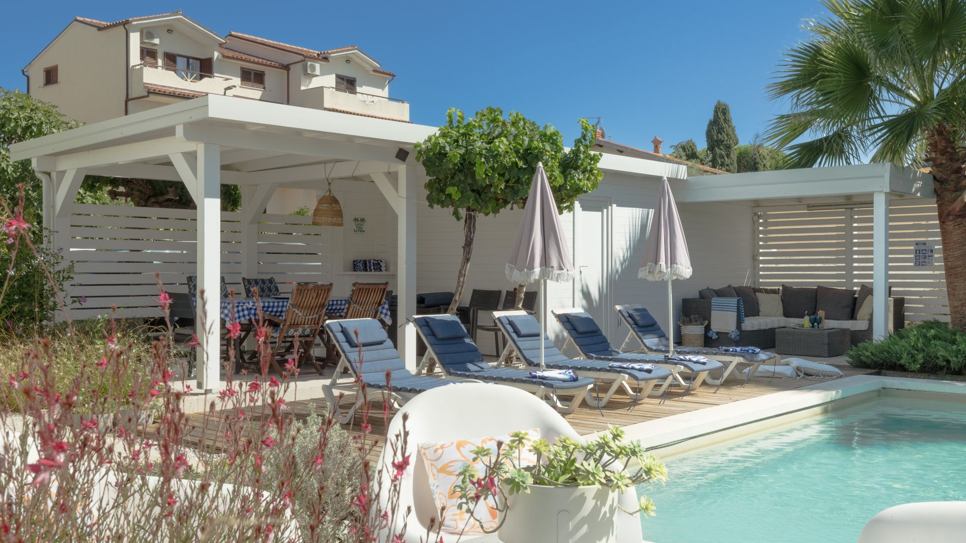 Un espace aménagé au bord de la piscine, avec des chaises longues, un coin salon abrité et un bâtiment en arrière-plan.