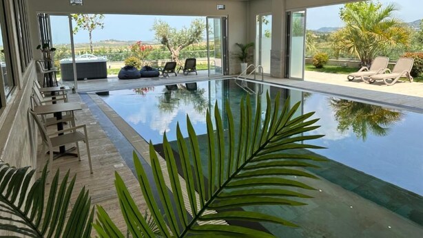 A modern indoor swimming pool with clear glass walls, lounge chairs, and a view of the landscape. - Villas by Top Villas (Maminas)
