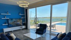 A modern living room with a large TV, blue sofas, and a view of a pool and water body. - Villas by Top Villas (Maminas)
