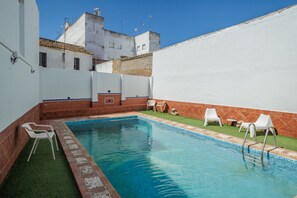 Piscina com borda azulejada, cercada por paredes brancas e uma pequena área gramada com cadeiras brancas.