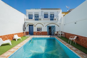 Uma piscina com terraço solar, paredes brancas e um edifício de dois andares com varandas.
