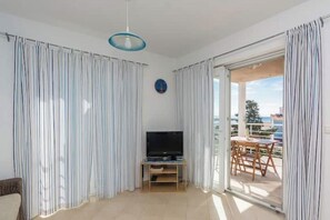 A room with a TV on a wooden stand, a ceiling light, and a view of a balcony with outdoor furniture. - Rogina 1 apartment (Novalja)