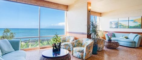 A spacious living area with a large window overlooking the ocean, a blue sofa, patterned armchairs, and a round coffee table.