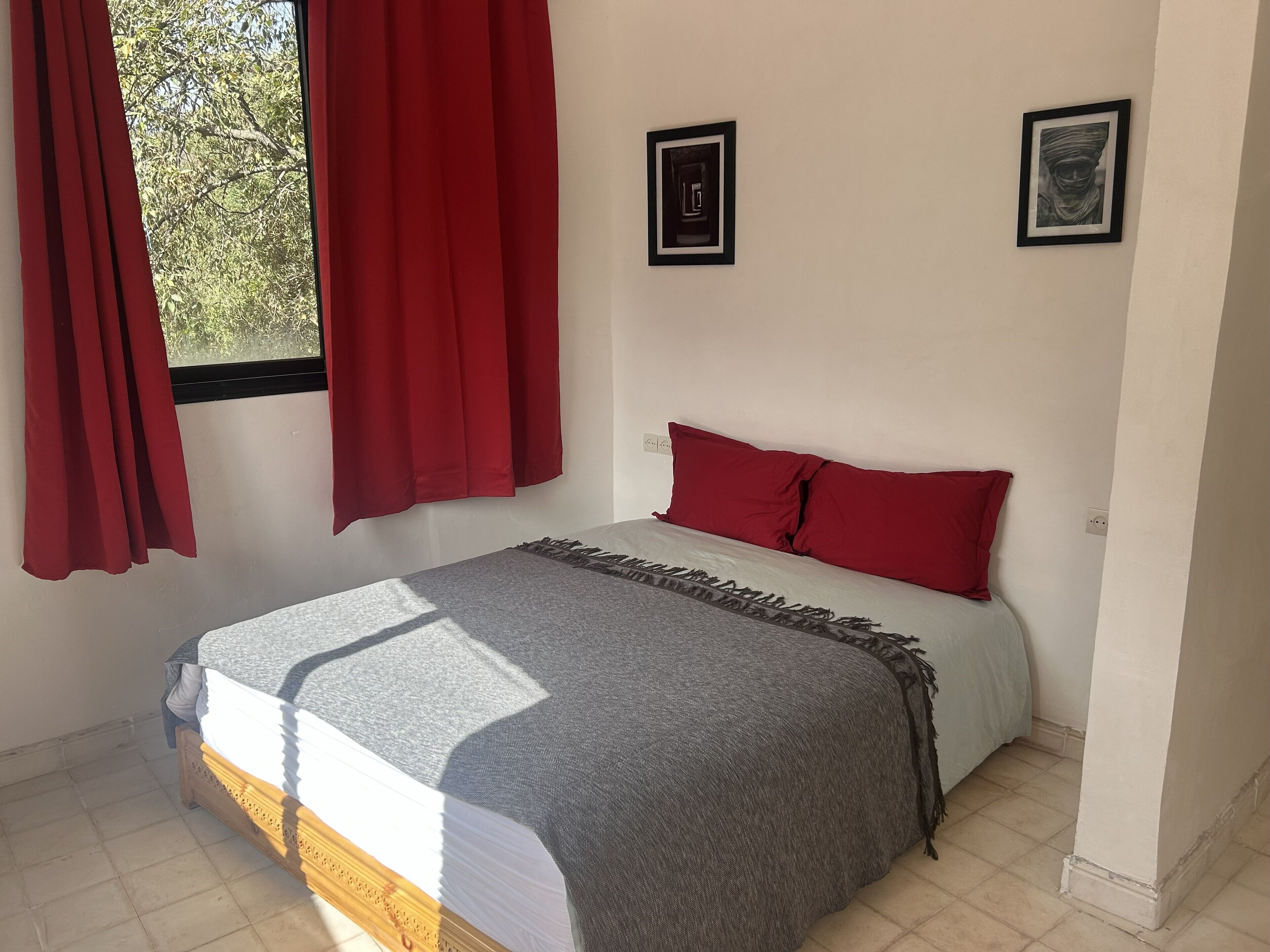 Ein Schlafzimmer mit einem Bett, roten Vorhängen, zwei gerahmten Bildern an der Wand und einem Fenster mit roten Vorhängen.