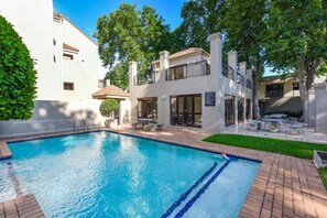 Piscina com água azul cristalina, cercada por um terraço de tijolos e um edifício branco com varanda.