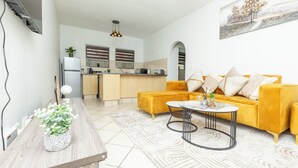 Un salon moderne avec un canapé jaune, une table basse et une cuisine en arrière-plan.